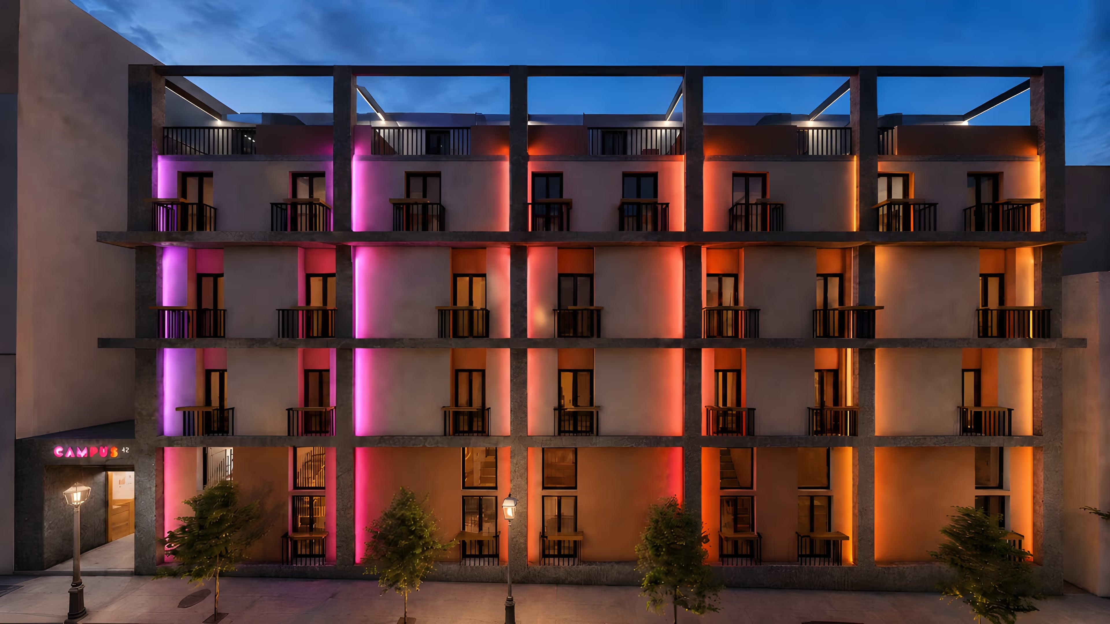 Campus42 façade at dusk — magenta and orange brand lighting washes the four-storey concrete frame on Georgiou Olympiou 42, Patras.